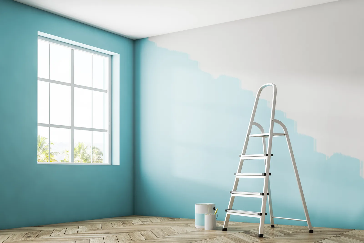 A bright, partially painted room with soft blue walls, where the upper portion remains white. A silver step ladder stands against the wall beside an open paint can on a light wooden floor. Sunlight streams through a large window, illuminating the space and highlighting the fresh paint.